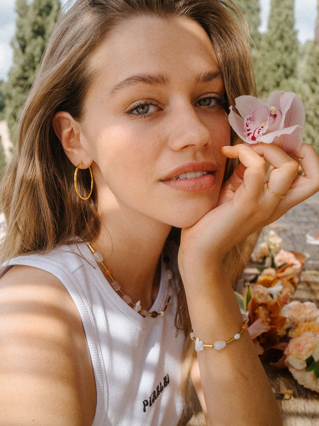 Zarte PURELEI Halskette und Armband mit Muscheln, getragen in warmem Licht, umgeben von Blumen.