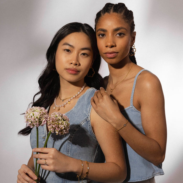 Zarte PURELEI Schmuckstücke, Perlenkette und goldene Creolen, umgeben von floraler Leichtigkeit.