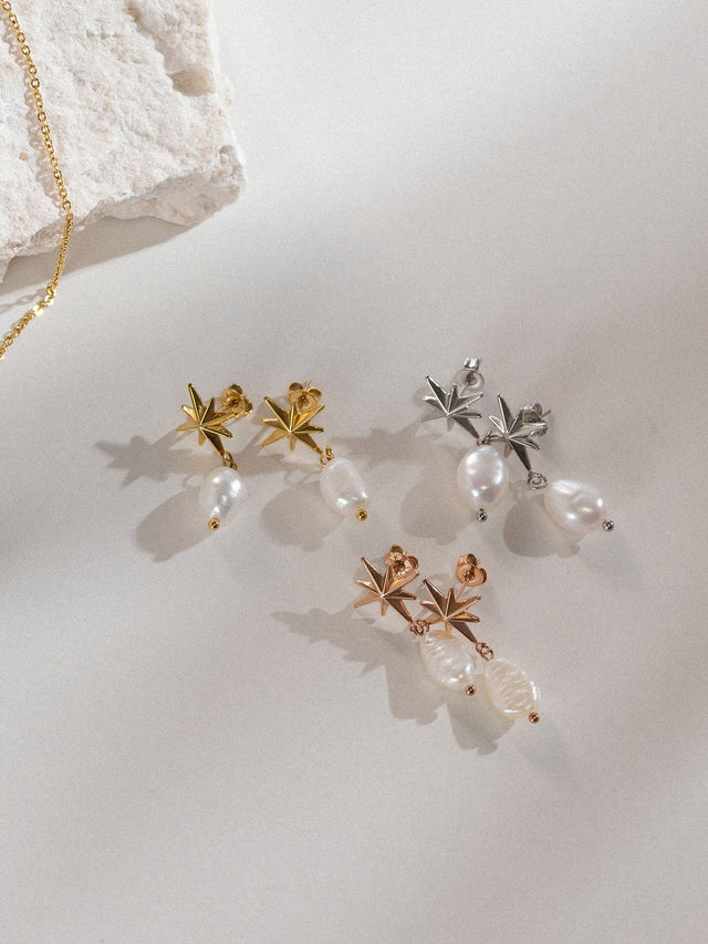 Mahina Club Windrose Ohrringe in Gold, Silber und Roségold mit schimmernder Süßwasserperle, elegant inszeniert auf hellem Untergrund.