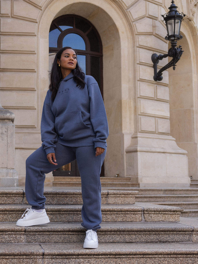 Bequemer, blauer Hoodie und Jogginghose von PURELEI, lässig vor eleganter Architektur.
