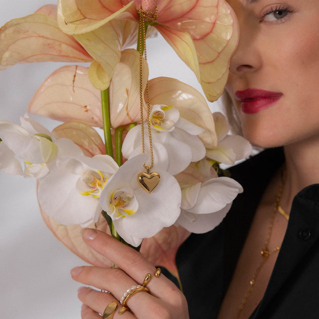 Goldene Herz-Kette von PURELEI, umgeben von weißen Orchideenblüten, elegant und feminin.