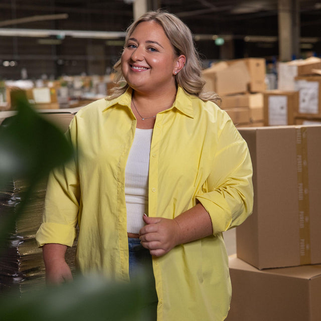 Frau in gelber Bluse und weißem Top, lächelt vor Kartons, PURELEI inspiriert.