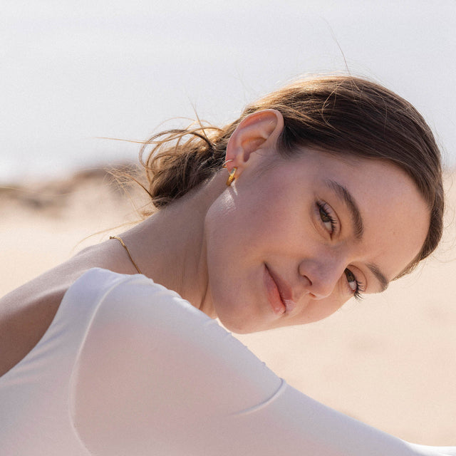 Elegante goldene Ohrringe von PURELEI, getragen am Strand, strahlen feminine Leichtigkeit aus.