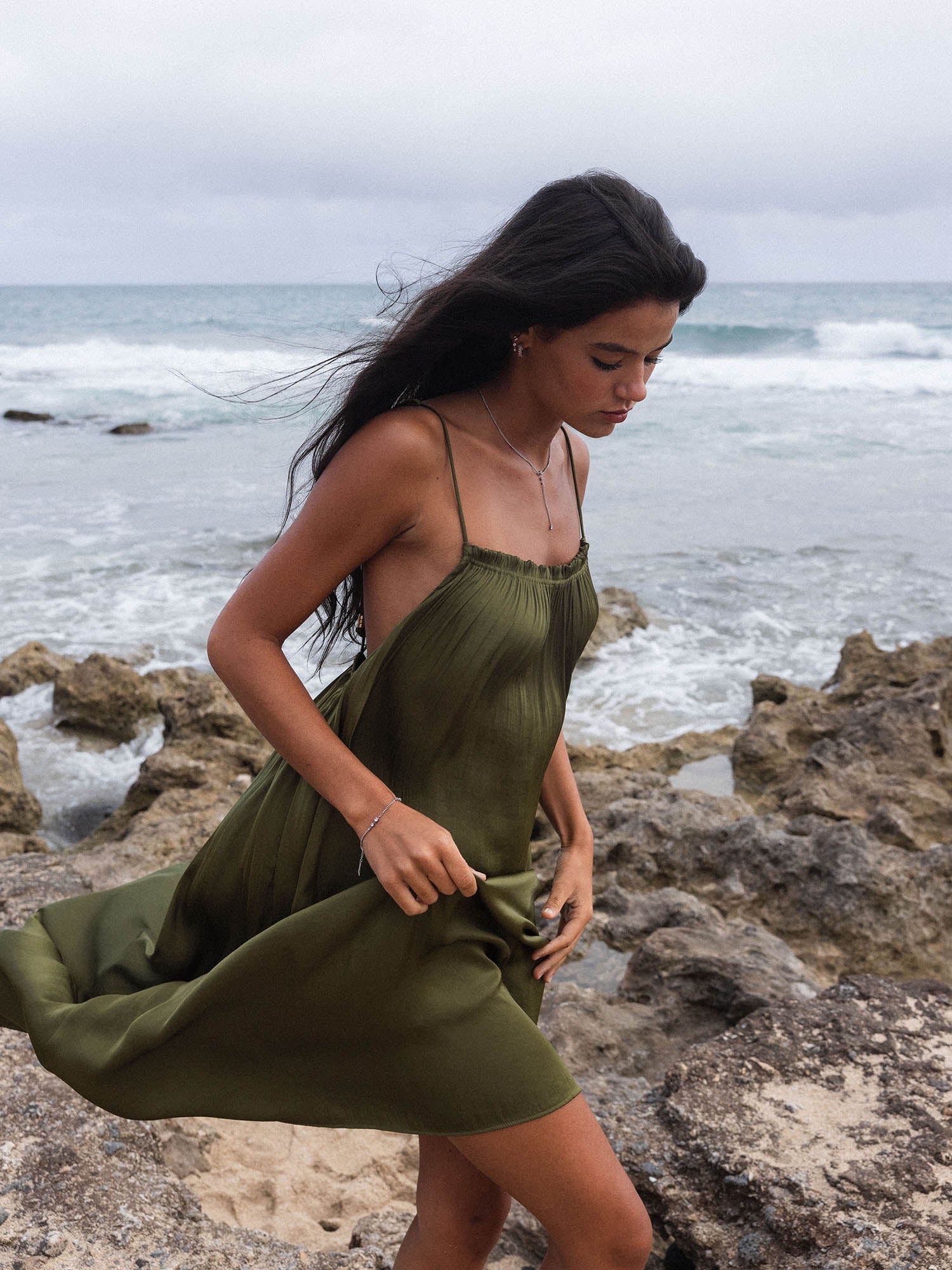Braunes, luftiges Kleid am Strand mit PURELEI Schmuck, umgeben von Wellen und Felsen.