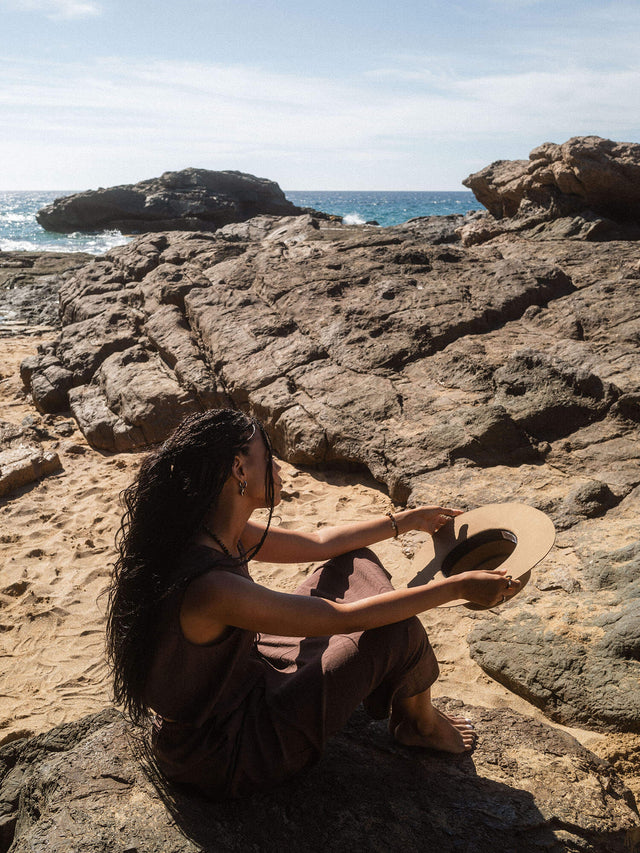 Frau mit Hut sitzt auf Felsen am Strand, PURELEI inspiriert von tropischer Eleganz.