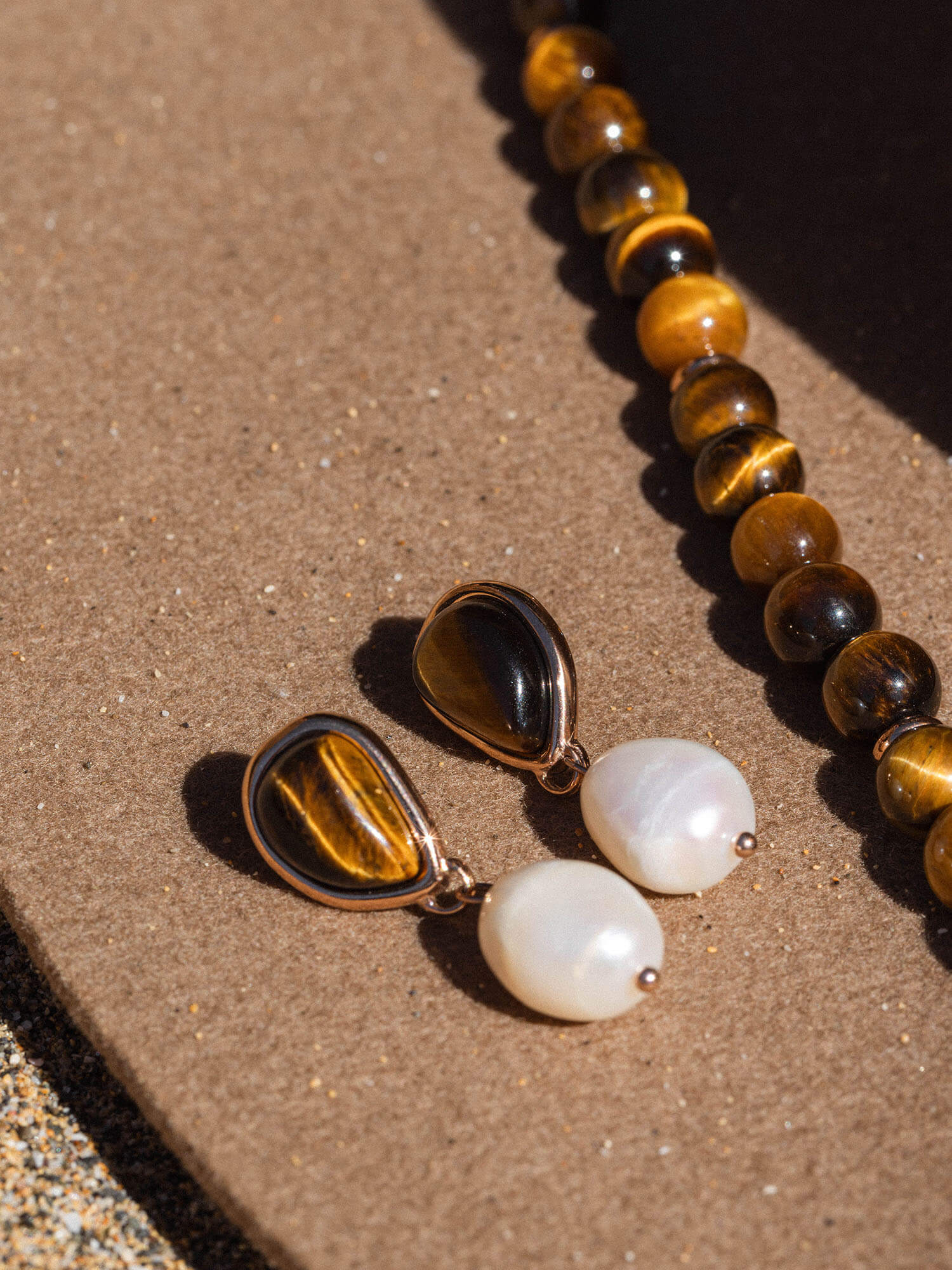 Zarte Ohrringe mit Tigerauge und weißen Details, PURELEI, auf sandfarbenem Untergrund.
