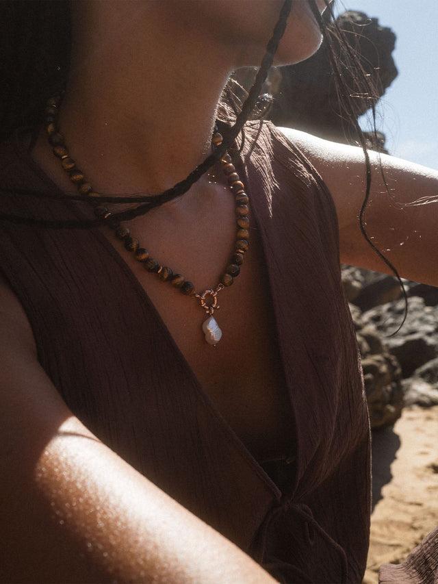 Braune Perlenkette mit Muschel-Anhänger, getragen in sonniger Strandatmosphäre – PURELEI.