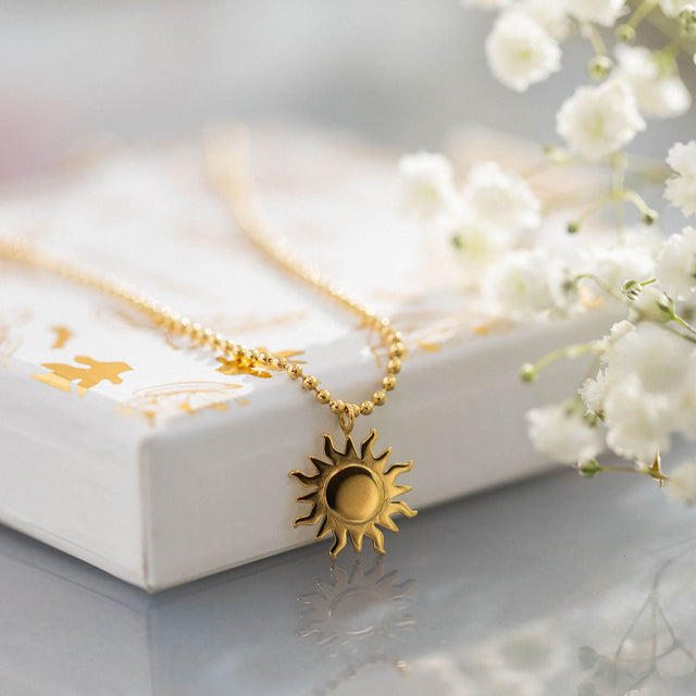 Goldene PURELEI Sonnensonnenkette auf hellem Untergrund, umgeben von zarten Blumen.