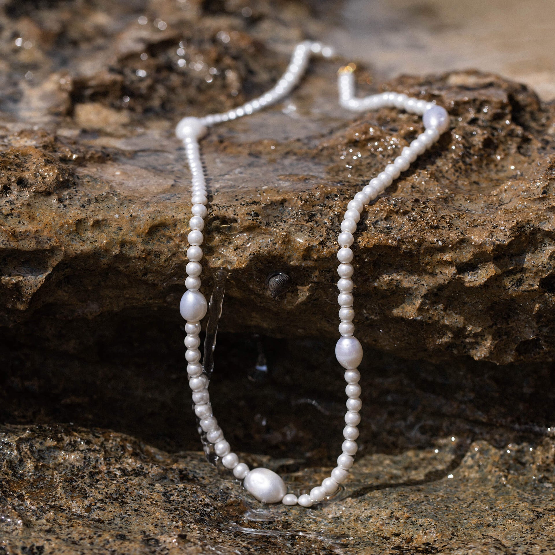 PURELEI Glace Kette mit Süßwasser- und Muschelperlen auf nassem Felsen, feminine Eleganz und Insel-Spirit.