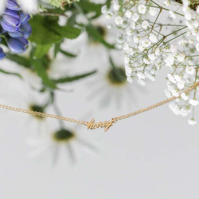 Honey Kette von PURELEI in 18K Gold mit filigranem Schriftzug, elegant inszeniert mit zarten Blüten.