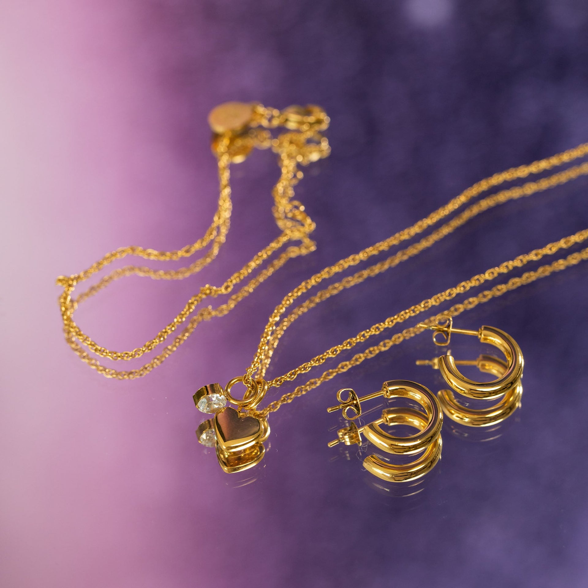 Goldene Kette mit Herz-Anhänger und Creolen von PURELEI auf lila Hintergrund.
