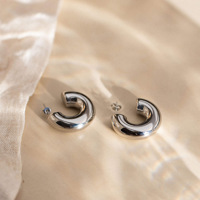 Silberne Creolen von PURELEI mit geschwungenem Design, elegant auf sanftem Stoff inszeniert.